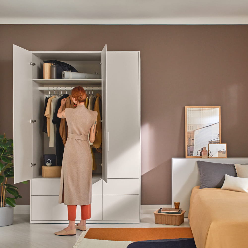 A three door Cashmere Beige Wardrobe with hanging rack, shelf and external drawers stands against a grey bedroom wall.