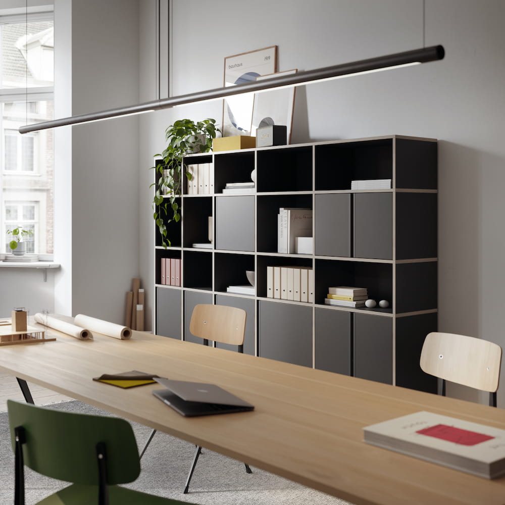 A minimalist black Type01 Wall Storage stands against a grey wall in a modern office with four rows of mixed open and closed sections displaying and concealing office supplies, books and more.