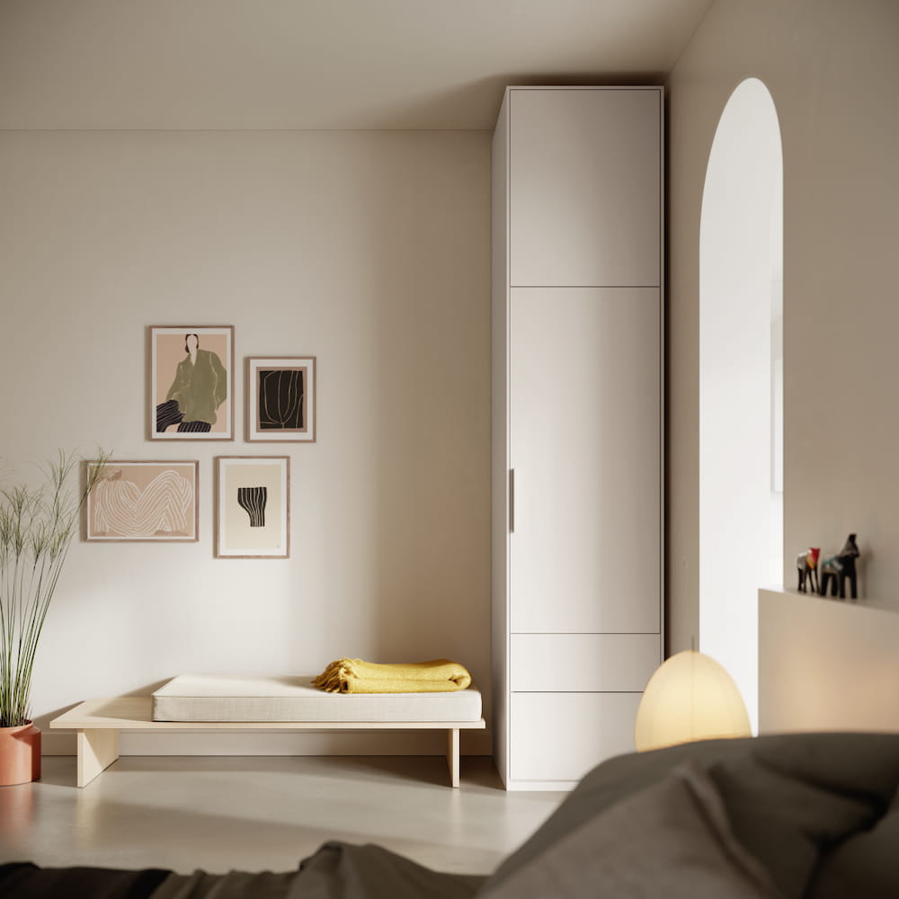 A tall, slim one-door Tone Wardrobe in white with top storage and one external drawer stands snugly in a narrow corner of a white bedroom.